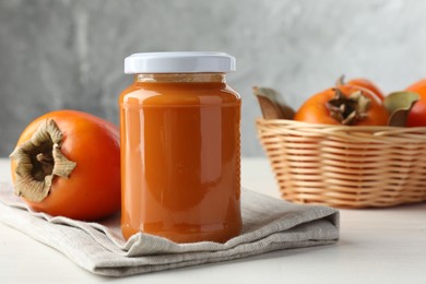 Photo of Delicious persimmon jam in glass jar and fresh fruit on white wooden table