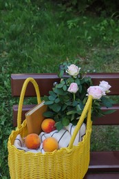 Yellow wicker bag with roses, book and peaches on bench outdoors