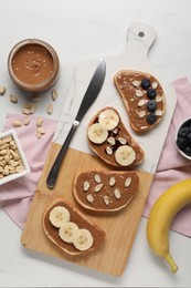 Photo of Toasts with tasty nut butter, banana slices, blueberries and peanuts on white table, flat lay