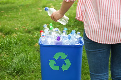 Photo of Woman throwing used bottle into trash bin outdoors, closeup. Plastic recycling