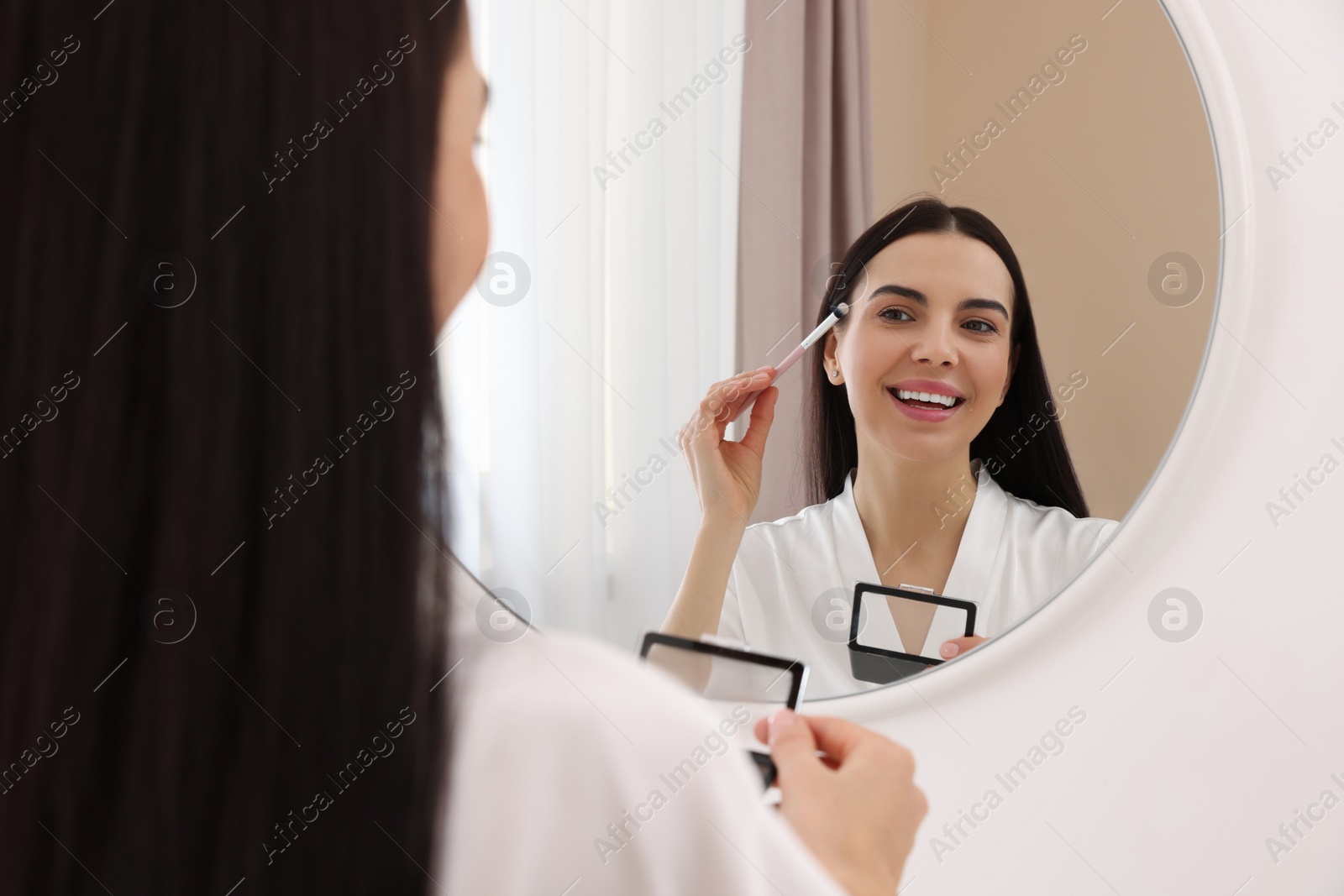 Photo of Beautiful young woman applying eyeshadow with brush near mirror indoors