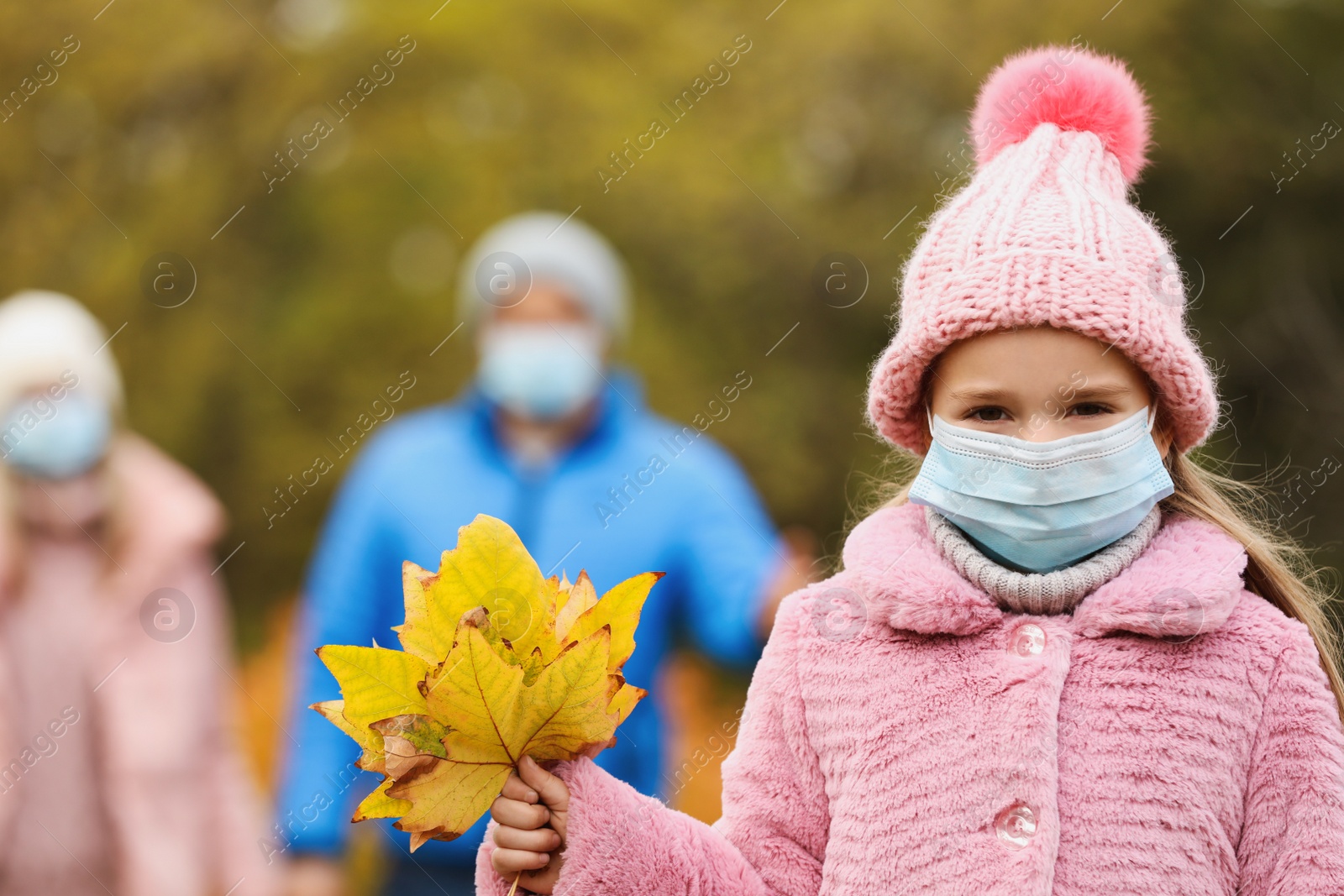 Photo of Cute girl in medical mask walking with her parents outdoors on autumn day. Protective measures during coronavirus quarantine