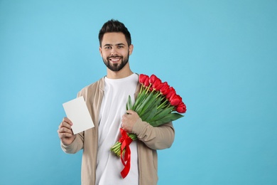 Photo of Happy man with red tulip bouquet and greeting card on light blue background, space for text. 8th of March celebration
