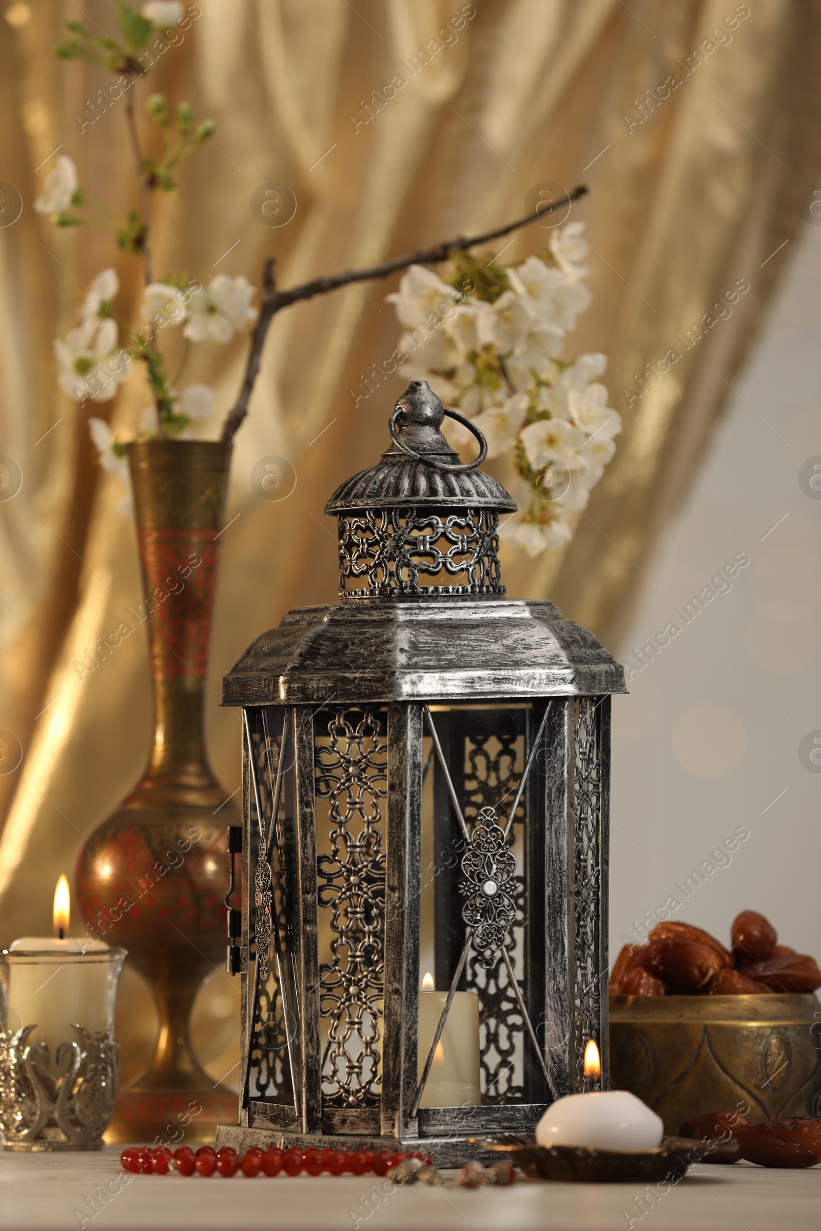 Photo of Arabic lantern, misbaha, candles, dates and flowers on table against blurred lights