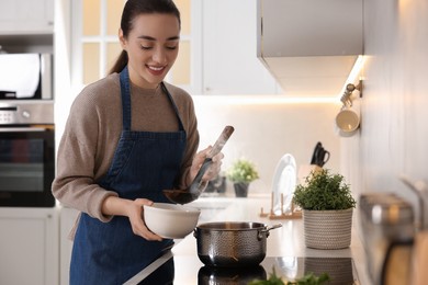 Photo of Smiling woman pouring tasty soup into bowl at countertop in kitchen