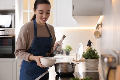 Smiling woman pouring tasty soup into bowl at countertop in kitchen