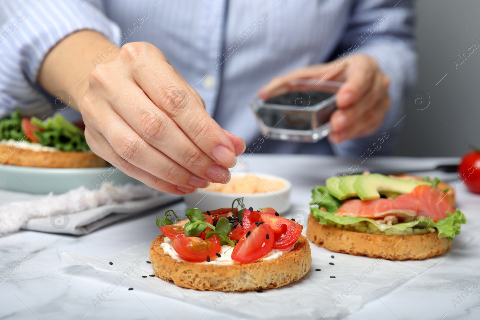 Photo of Woman adding black sesame seeds onto tasty rusks with different toppings at white marble table, closeup