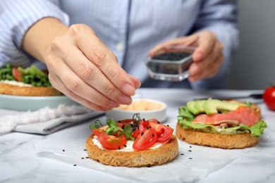 Woman adding black sesame seeds onto tasty rusks with different toppings at white marble table, closeup