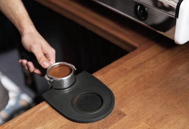 Photo of Barista holding portafilter with milled coffee on tamp mat at bar counter, closeup. Space for text