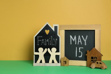 Photo of Happy International Family Day. Small chalkboards with text, house models, people figures and toy car on green table against yellow background