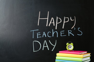 Photo of Books and alarm clock near blackboard with inscription HAPPY TEACHER'S DAY