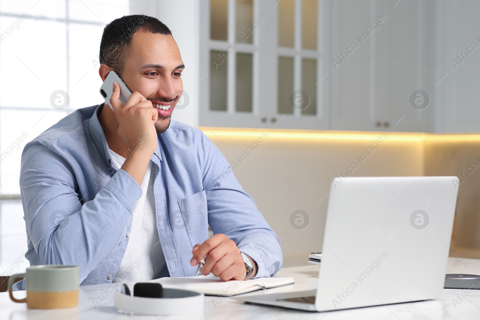 Photo of Young man talking on smartphone while working with laptop at desk in kitchen. Home office