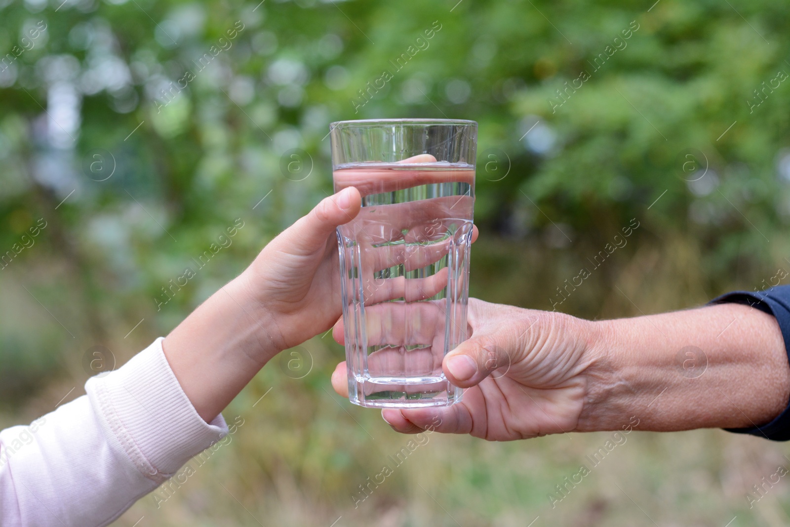 Photo of Child giving glass of water to elderly woman outdoors, closeup