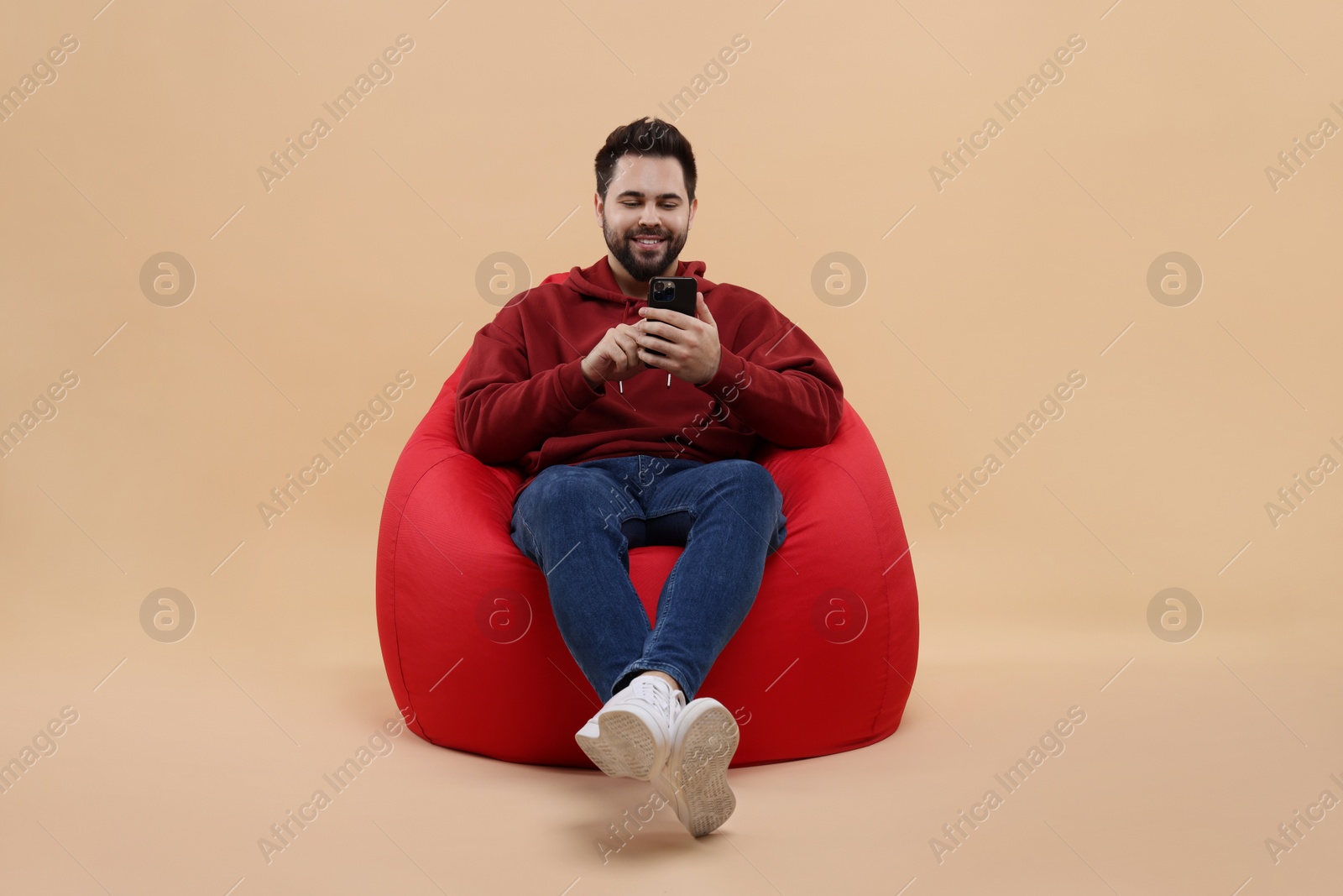 Photo of Happy young man using smartphone on bean bag chair against beige background