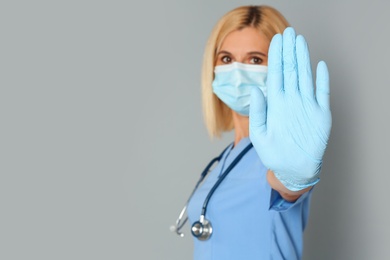 Photo of Doctor in protective mask showing stop gesture on grey background, space for text. Prevent spreading of coronavirus