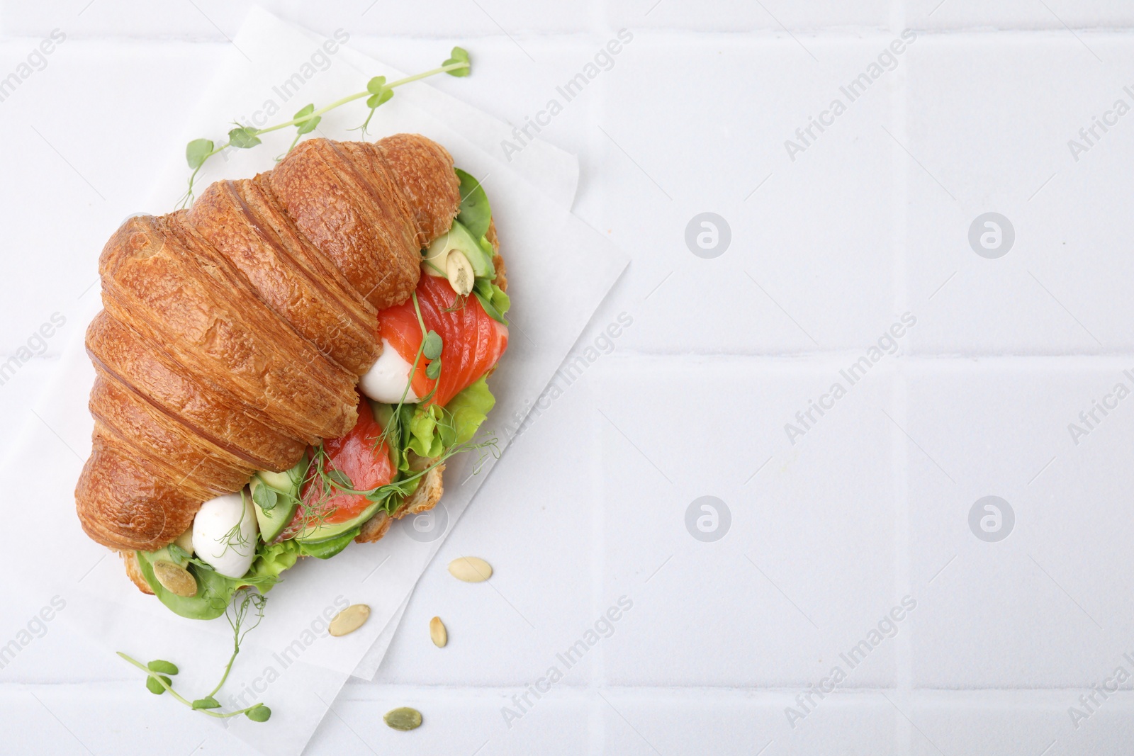 Photo of Tasty croissant with salmon, avocado, mozzarella and lettuce on white tiled table, top view. Space for text
