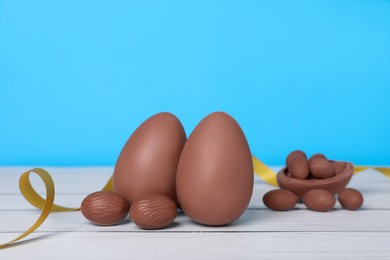 Photo of Delicious chocolate eggs and golden ribbon on white wooden table against light blue background, closeup
