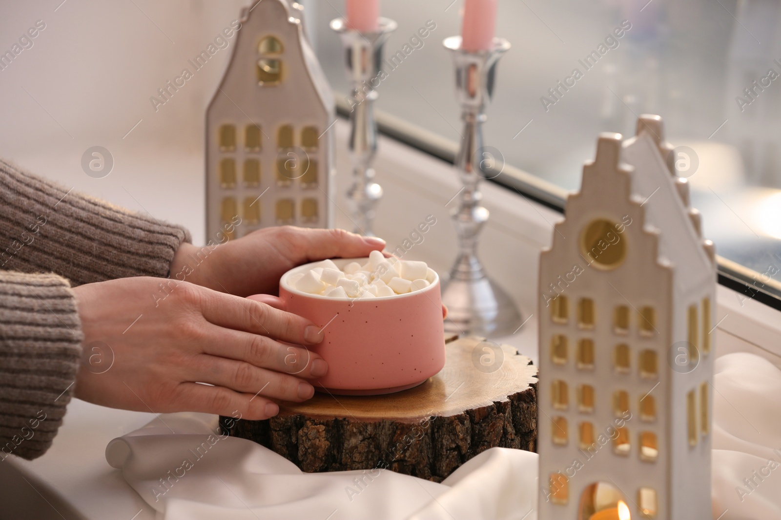 Photo of Woman holding cup of delicious drink near windowsill with house shaped lanterns indoors, closeup