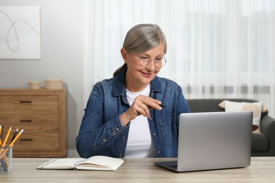 Photo of Beautiful senior woman with pen using laptop at wooden table indoors