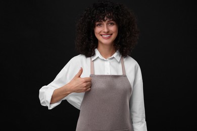 Happy woman wearing kitchen apron and showing thumbs up on black background. Mockup for design