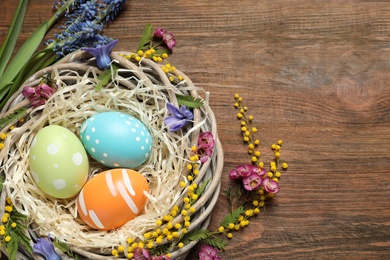 Flat lay composition of wicker nest with painted Easter eggs and flowers on wooden table, space for text