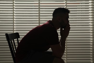 Photo of Sad man sitting near closed blinds indoors