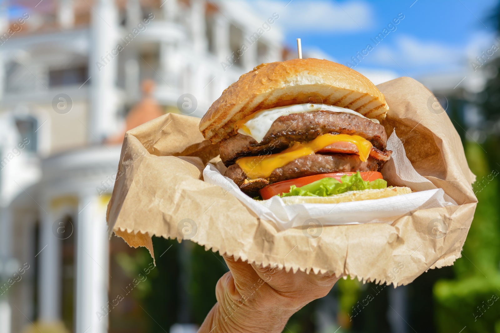 Photo of Woman holding delicious burger in paper wrap outdoors, closeup