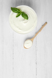 Photo of Bowl of fresh sour cream with basil and spoon on white wooden table, flat lay. Space for text