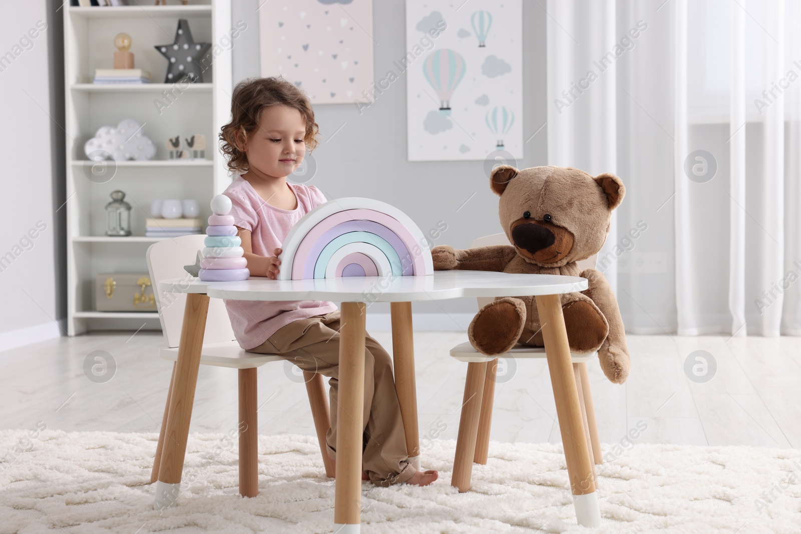 Photo of Cute little girl playing with toy and teddy bear at white table in room