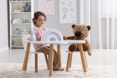 Cute little girl playing with toy and teddy bear at white table in room