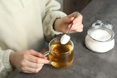 Woman adding sugar into aromatic tea at grey table, closeup