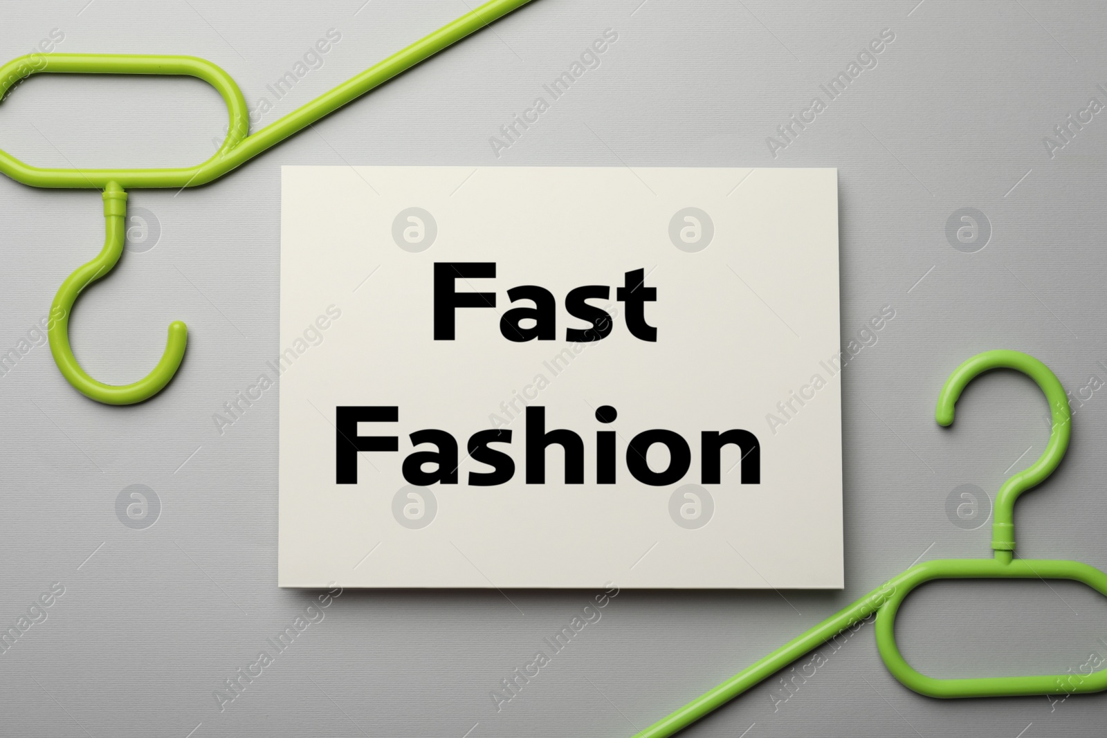 Photo of White card with phrase Fast Fashion and green hangers on light grey background, flat lay