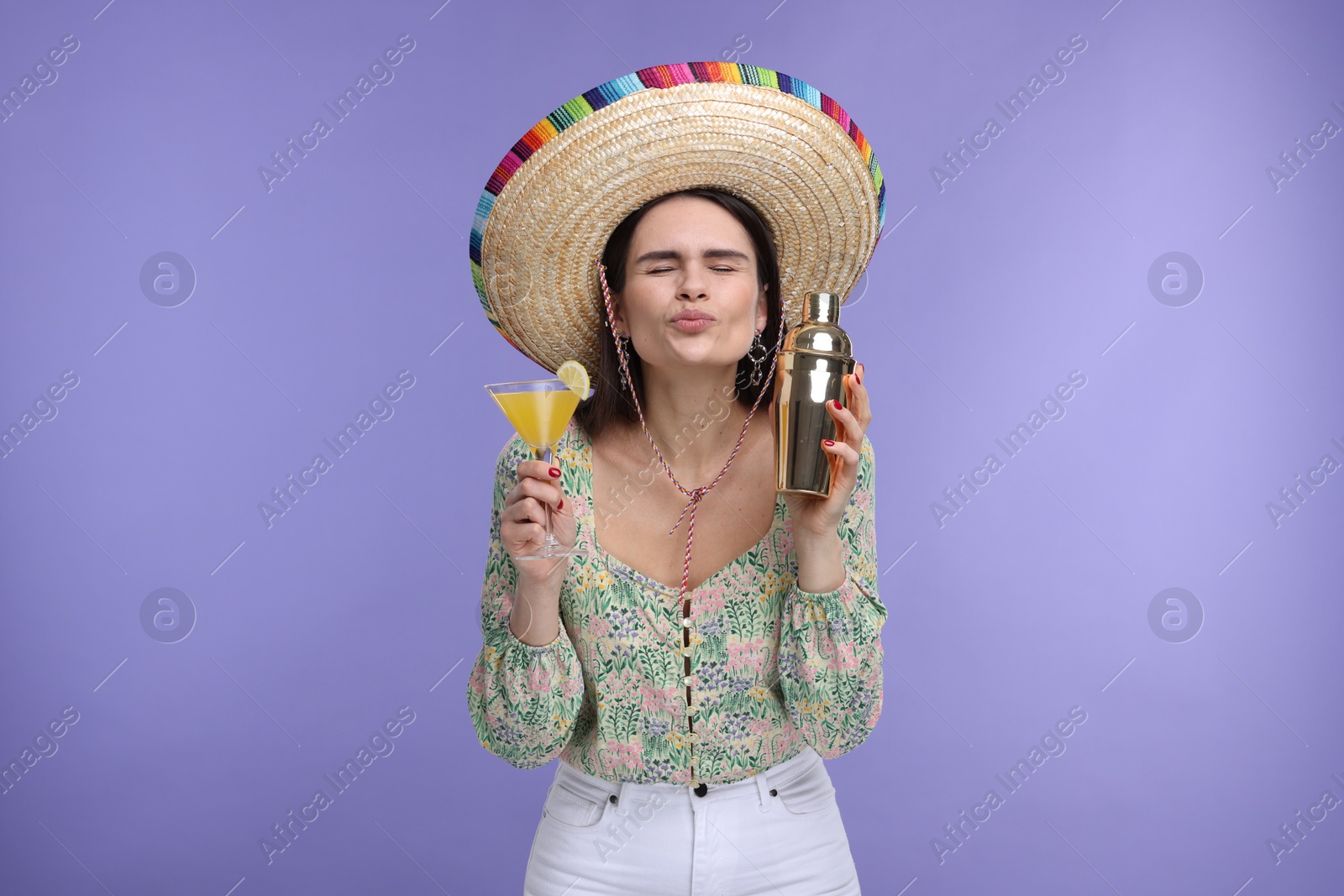 Photo of Young woman in Mexican sombrero hat with cocktail and shaker sending air kiss on violet background