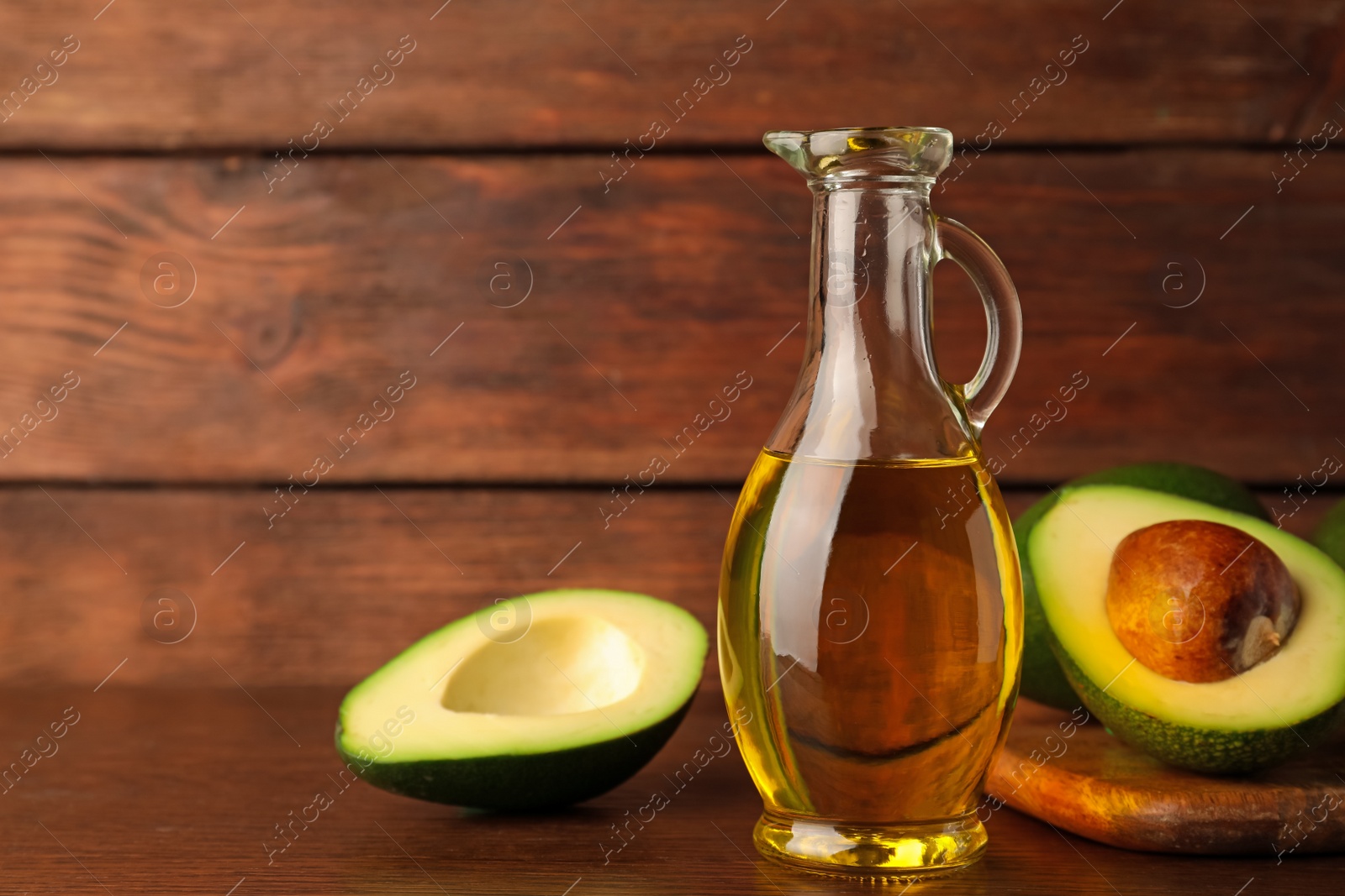 Photo of Glass jug of cooking oil and fresh avocados on wooden table, space for text