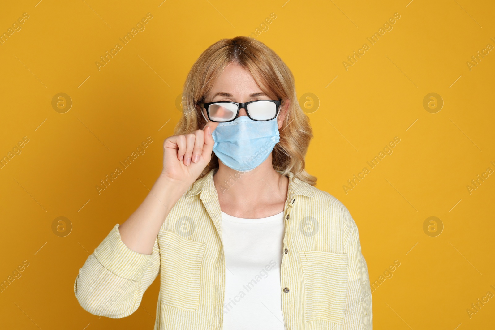 Photo of Woman wiping foggy glasses caused by wearing medical mask on yellow background