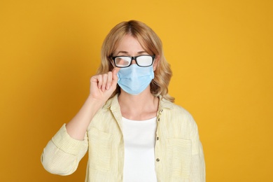 Woman wiping foggy glasses caused by wearing medical mask on yellow background