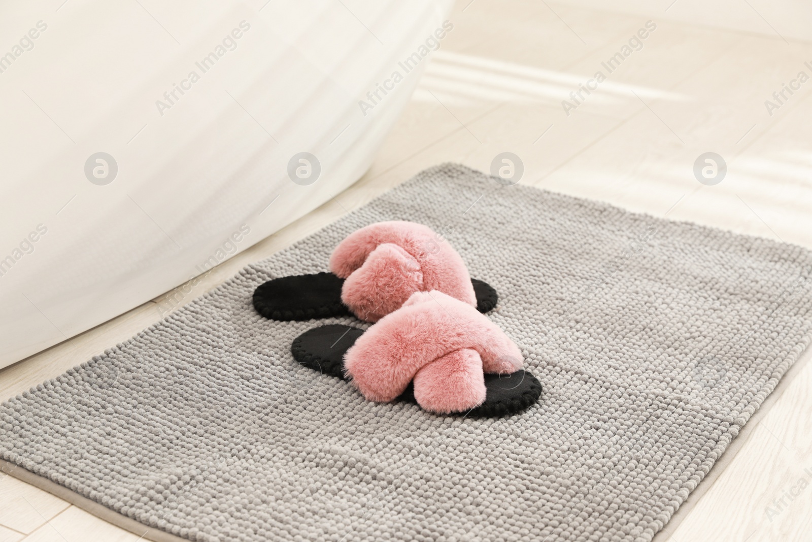 Photo of New grey bath mat with fluffy slippers near tub indoors