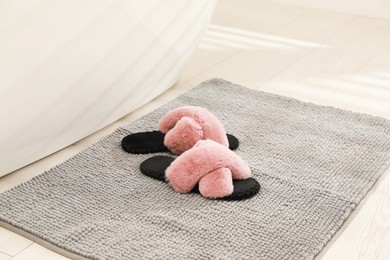 Photo of New grey bath mat with fluffy slippers near tub indoors