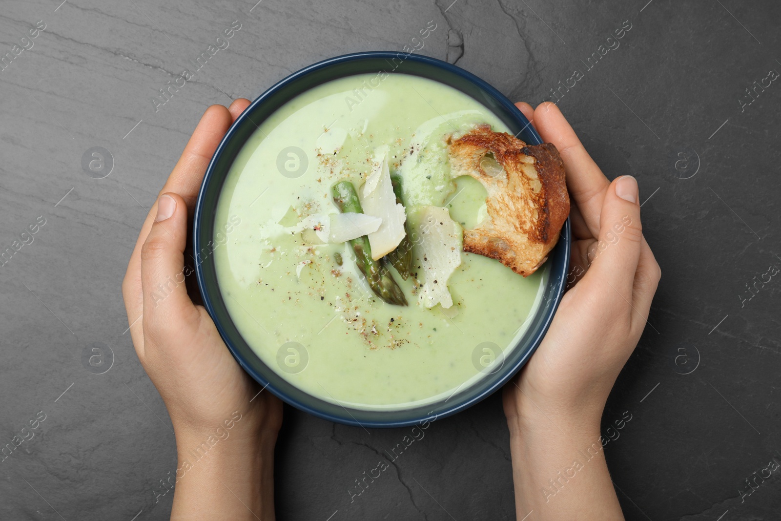 Photo of Woman with bowl of delicious asparagus soup at dark table, top view