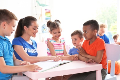 Photo of Young volunteer reading book with children at table indoors