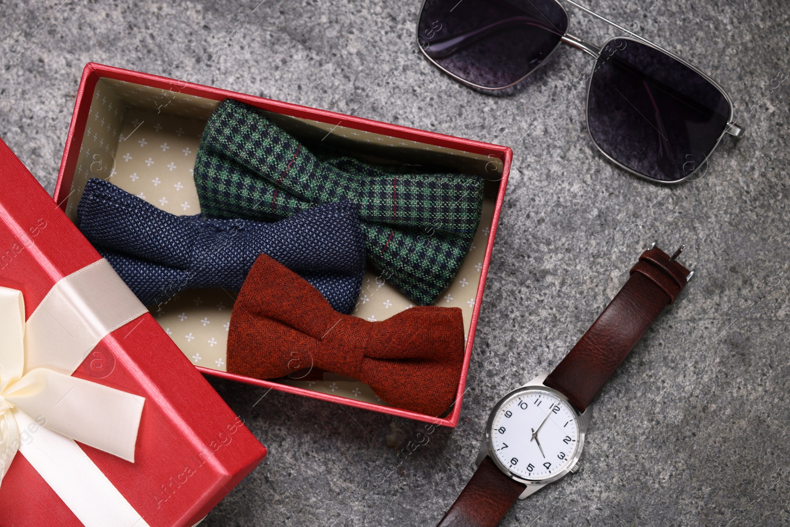 Photo of Stylish color bow ties in gift box, wristwatch and sunglasses on grey textured background, flat lay