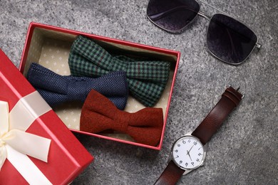 Photo of Stylish color bow ties in gift box, wristwatch and sunglasses on grey textured background, flat lay