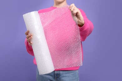 Woman holding roll of bubble wrap on purple background, closeup. Stress relief