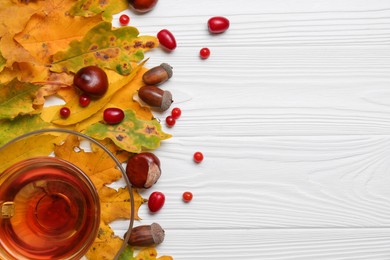 Different autumn leaves, acorns, chestnuts, red berries and cup of tasty tea on white wooden table, flat lay. Space for text