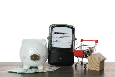 Electricity meter, house model, piggy bank and shopping cart with money on wooden table against white background