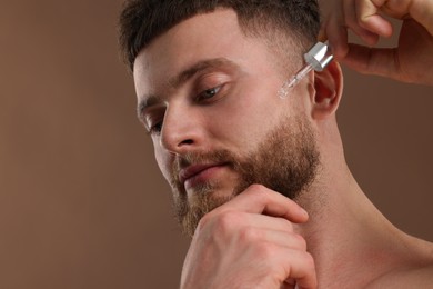 Handsome man applying serum onto his face on brown background, space for text