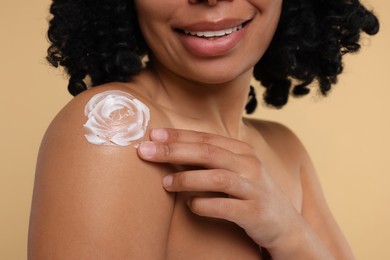 Young woman applying body cream onto shoulder on beige background, closeup