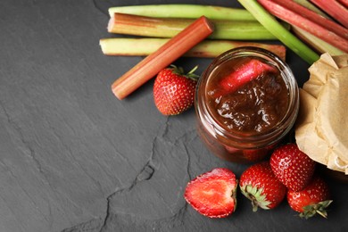 Photo of Jar of tasty rhubarb jam, fresh stems and strawberries on dark textured table, above view. Space for text