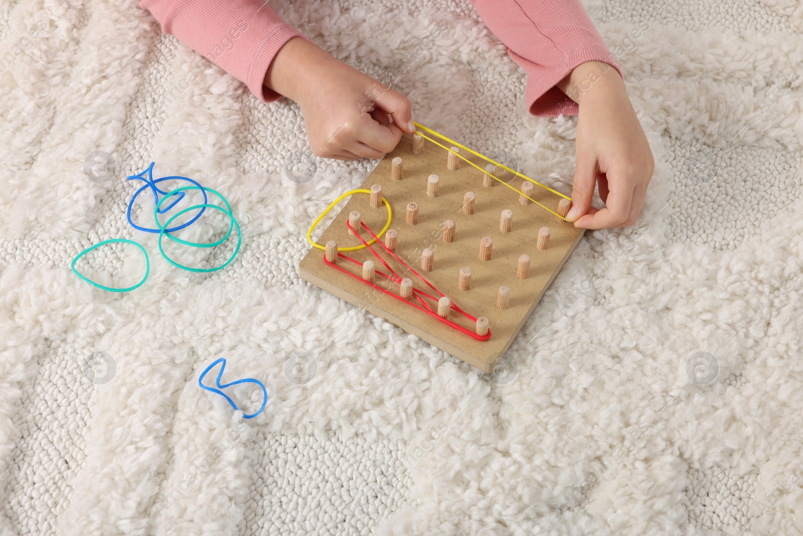 Photo of Motor skills development. Girl playing with geoboard and rubber bands on carpet, closeup