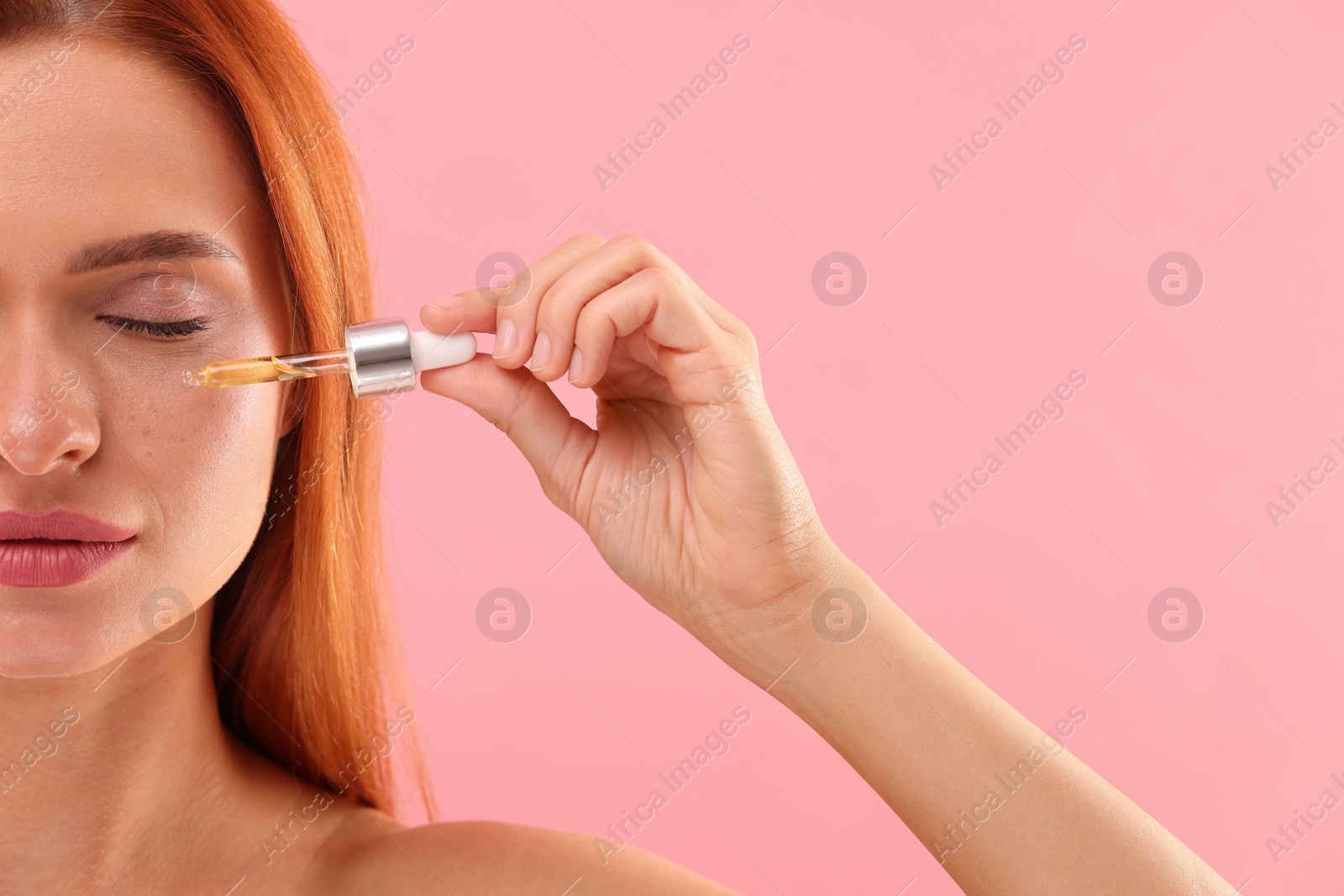 Photo of Beautiful young woman applying cosmetic serum onto her face on pink background, closeup. Space for text